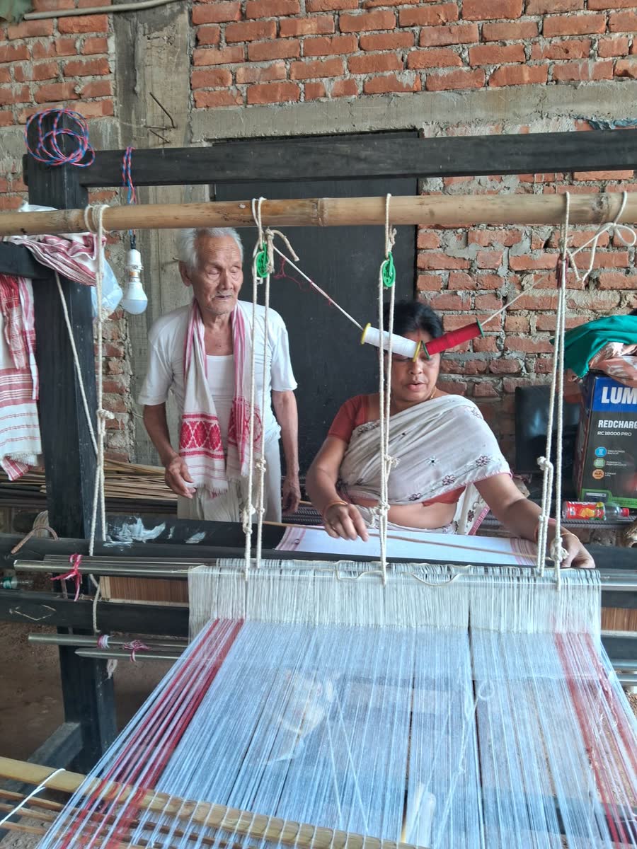 A woman weaving a gamosa on a traditional handloom while an elder looks on